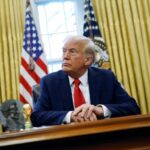A man in a suit sitting at a desk in the Oval Office with American flags in the background