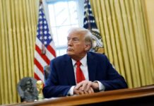 A man in a suit sitting at a desk in the Oval Office with American flags in the background